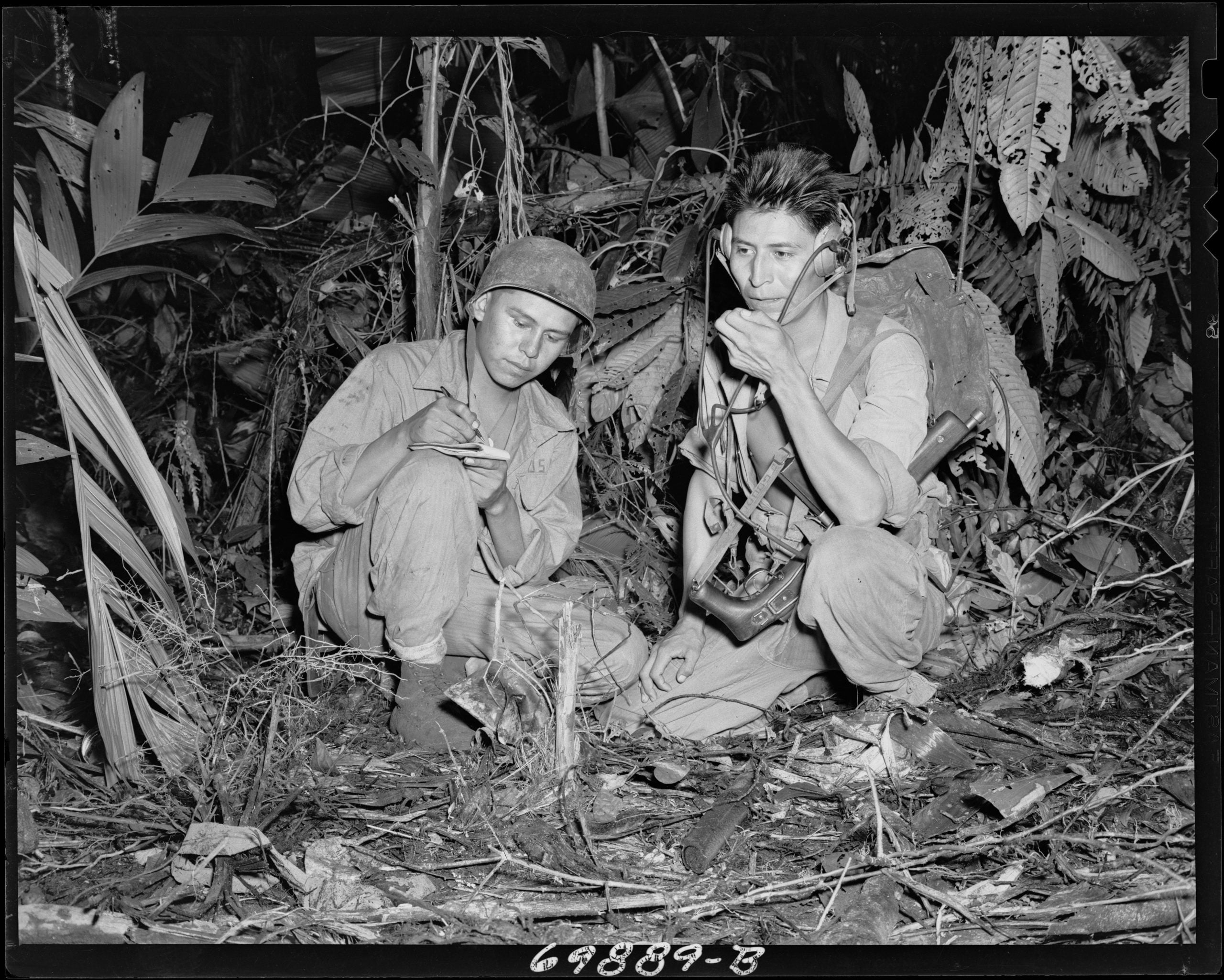 Two men sit in the jungle during WWII talking on a military radio